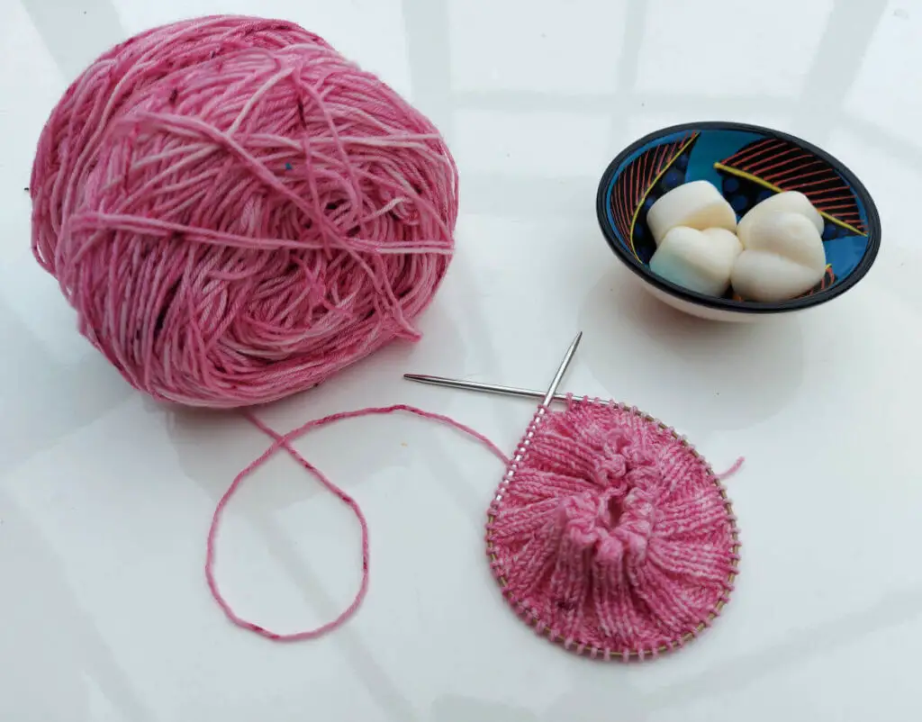 A partly-knitted pink sock is lying on a high gloss white table next to the ball of pink yarn and a small bowl of wax candle melts