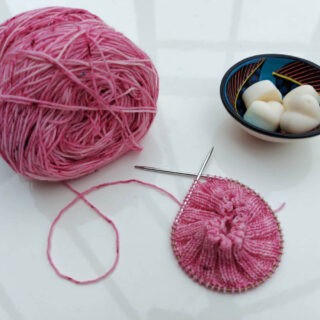 A partly-knitted pink sock is lying on a high gloss white table next to the ball of pink yarn and a small bowl of wax candle melts