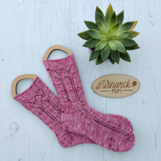 A pair of pink lacy socks on sock blockers lying on a white wooden board. Next to them is a wooden oval disk with the Winwick Mum logo engraved on it, and a green succulent plant