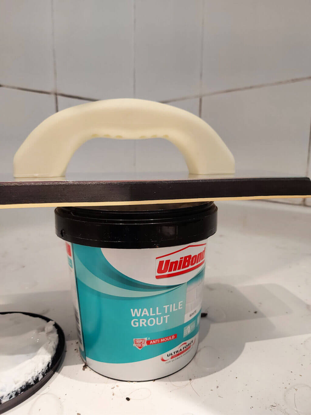 A tub of grout with a grout spreader balanced across it in a shower