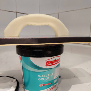 A tub of grout with a grout spreader balanced across it in a shower