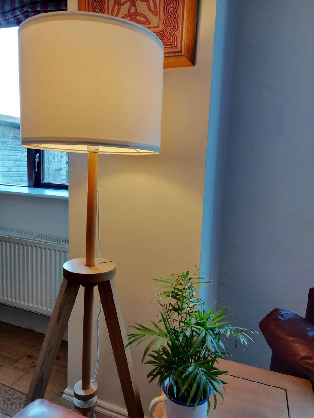A tall lamp on a three-legged stand is against a wall behind a sofa. There is a pink Celtic tapestry on the wall and a green leafy plant on the coffee table in front of it.