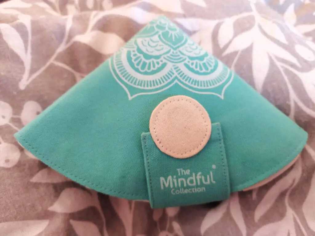 A triangular turquoise fabric case with a large cream circular closure is resting on a grey and white patterned duvet cover