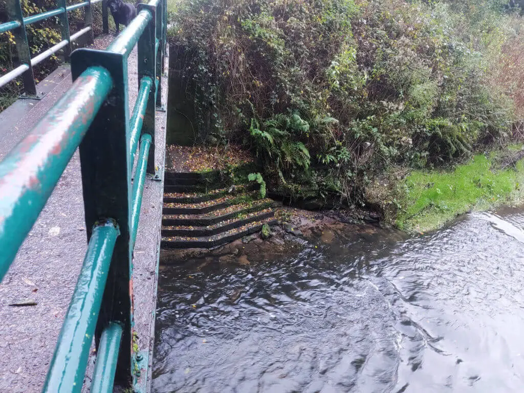 A bridge with green railings over a brook. There are steps under the bridge leading down to the water.