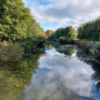 Looking out from a bridge across a canal. The cloudy sky is reflected in the water as are the trees lining the canal