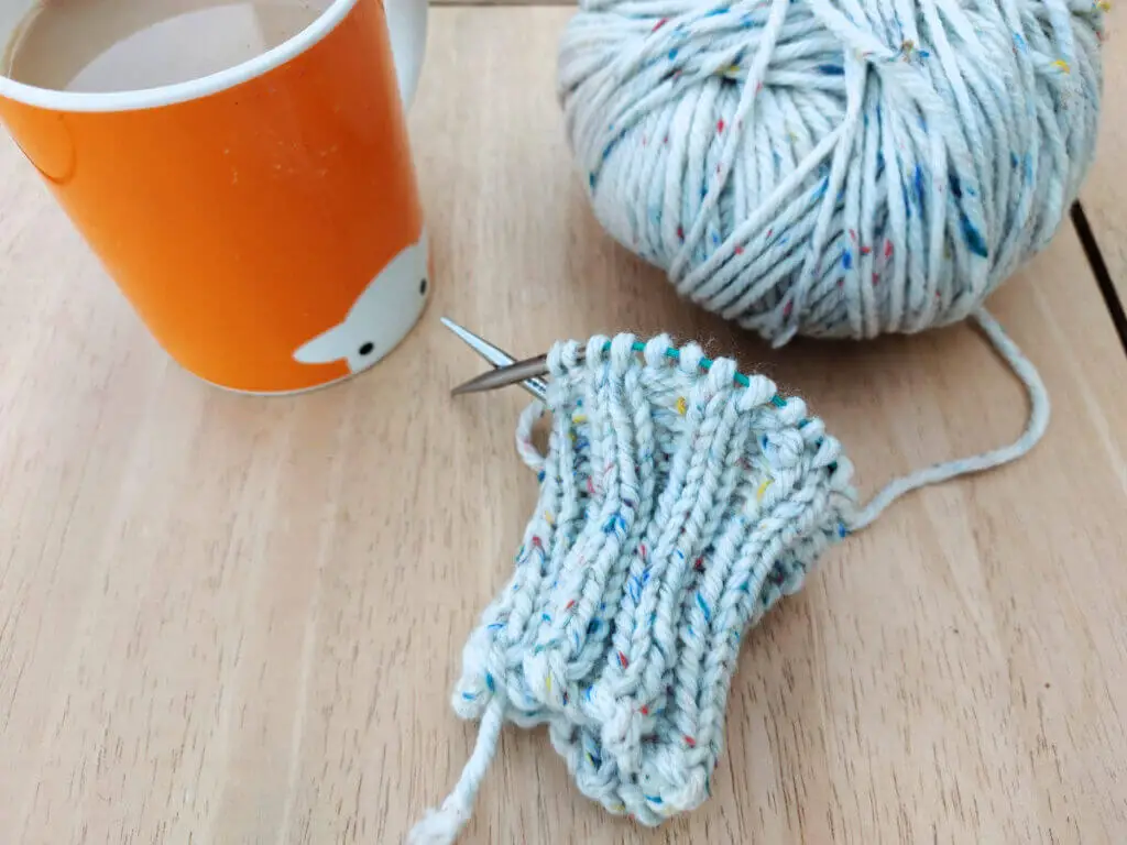 A knitted ribbed cuff on a short circular needle lying on a wooden table next to a ball of yarn and an orange mug of tea