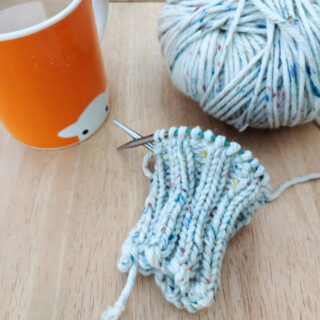A knitted ribbed cuff on a short circular needle lying on a wooden table next to a ball of yarn and an orange mug of tea