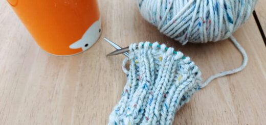 A knitted ribbed cuff on a short circular needle lying on a wooden table next to a ball of yarn and an orange mug of tea