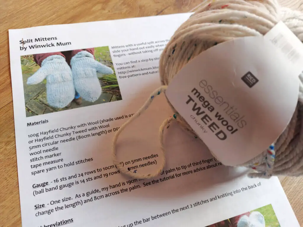 A ball of yarn sits on a paper pattern for Split Mittens, which is lying on a wooden floor