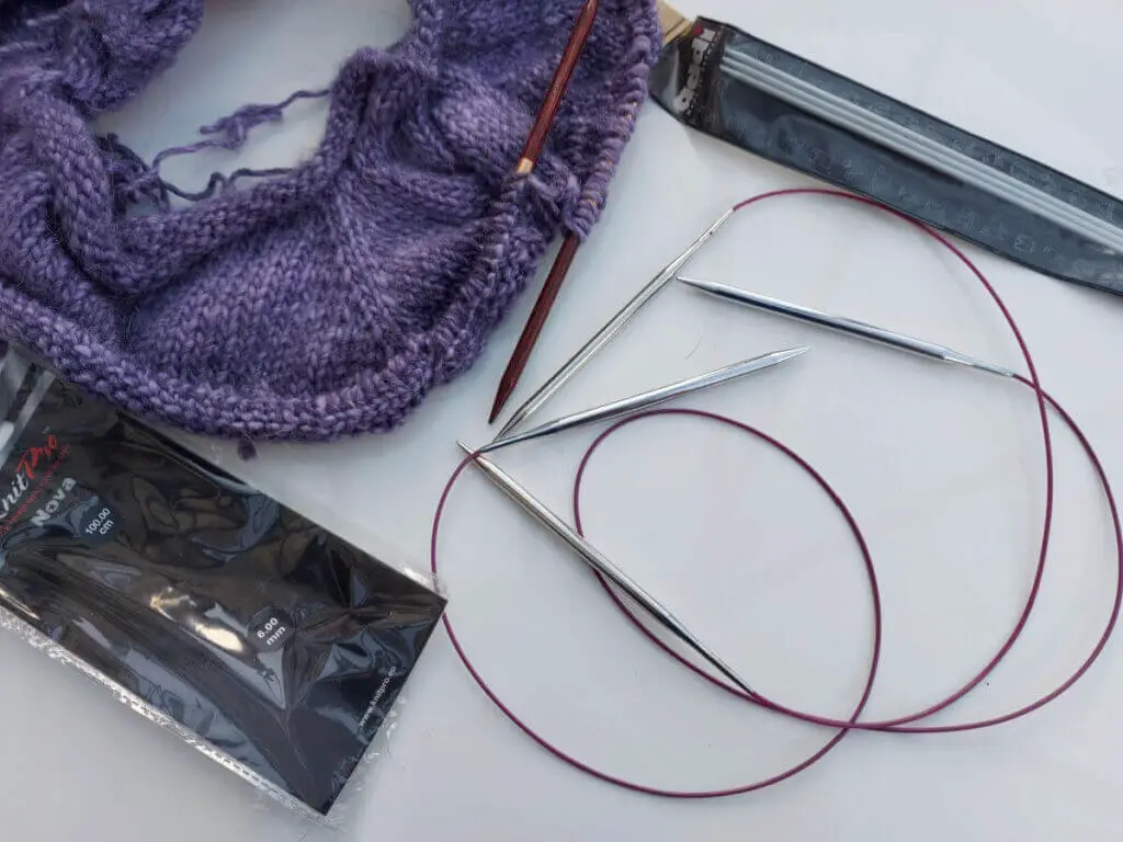 A partly-knitted purple jumper lies on a white high gloss table next to two long circular needles with metal needle tips, and a packet of three DPNs and an empty circular needle packet