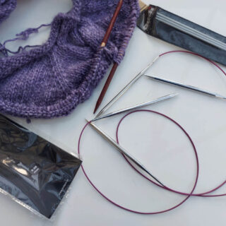 A partly-knitted purple jumper lies on a white high gloss table next to two long circular needles with metal needle tips, and a packet of three DPNs and an empty circular needle packet