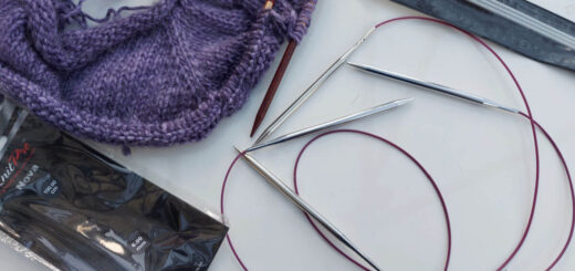 A partly-knitted purple jumper lies on a white high gloss table next to two long circular needles with metal needle tips, and a packet of three DPNs and an empty circular needle packet