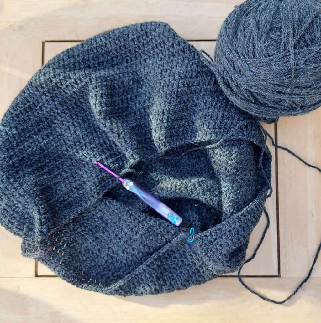 A grey crochet bag shape lies on a pale wooden table next to a large ball of grey yarn