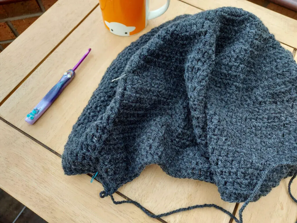 A grey crochet project is lying on a wooden table next to a crochet hook and an orange mug of tea