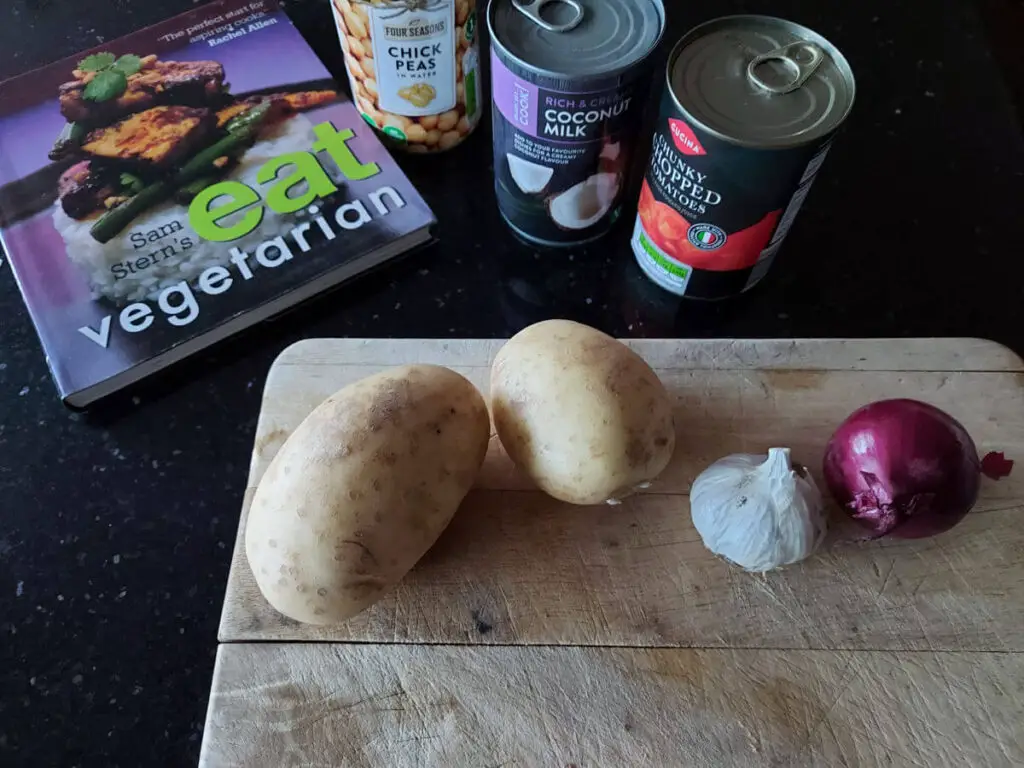A wooden chopping board on a black worktop. On the board are two potatoes, a red onion and a garlic clove. Next to the board are a can of tomatoes, a can of coconut milk and a can of chickpeas. A cookbook is next to them.