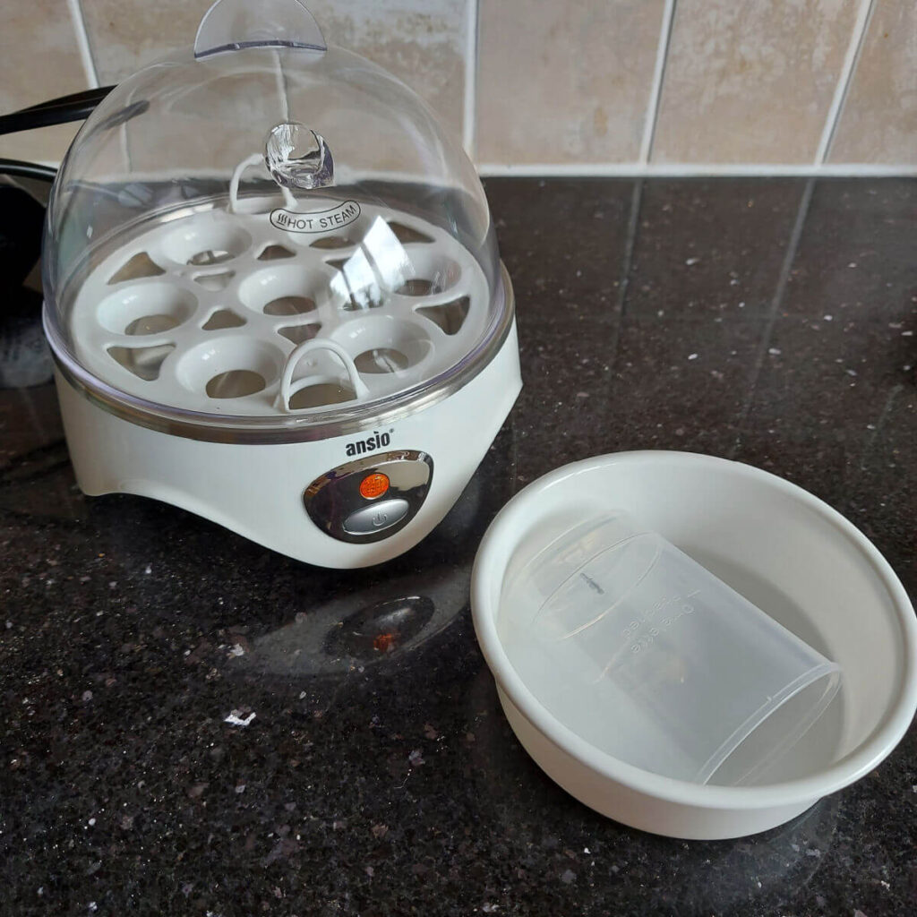 A white egg cooker with a clear lid on a black granite worktop. Next to it is a white bowl and a measuring jug