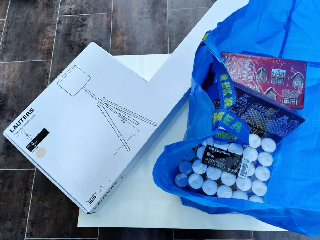 A blue IKEA bag containing tea lights, gingerbread house kits is on a white coffee table. There is a large box containing a lamp next to it.