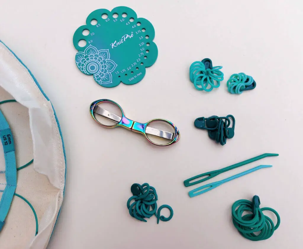 Turquoise plastic knitting accessories lie on a cream background. There is a needle gauge, two wool needles, a pair of scissors (they're not plastic) and five sets of stitch markers