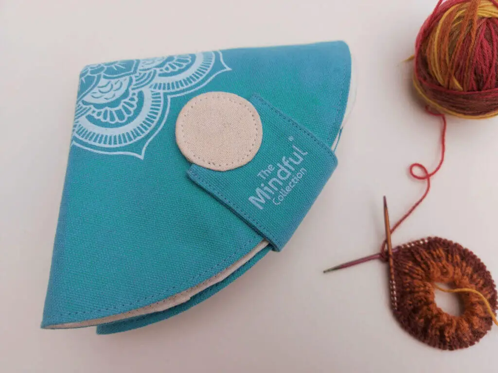 A triangular turquoise fabric case is lying on a cream background next to a half-knitted sock in shades of brown, red and yellow