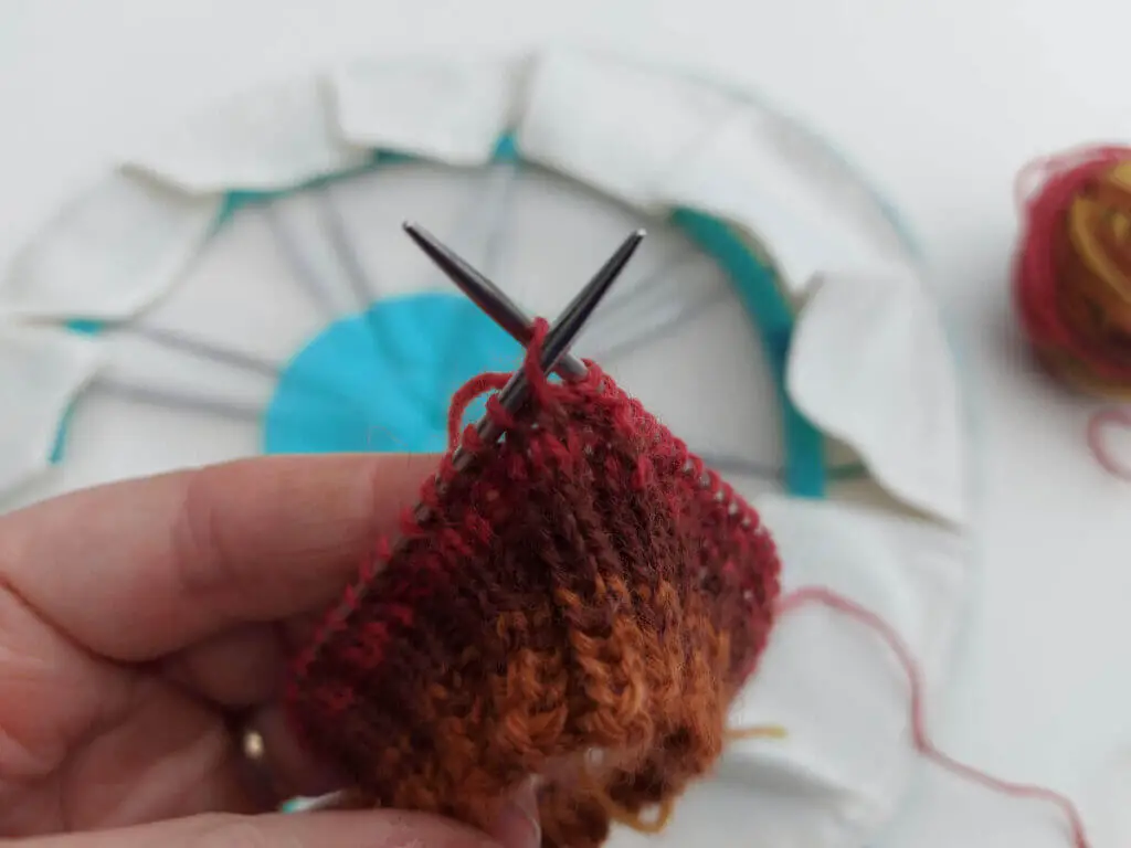 A sock cuff knitted in shades of brown, red and yellow on a metal short circular needle.