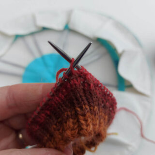 A sock cuff knitted in shades of brown, red and yellow on a metal short circular needle.