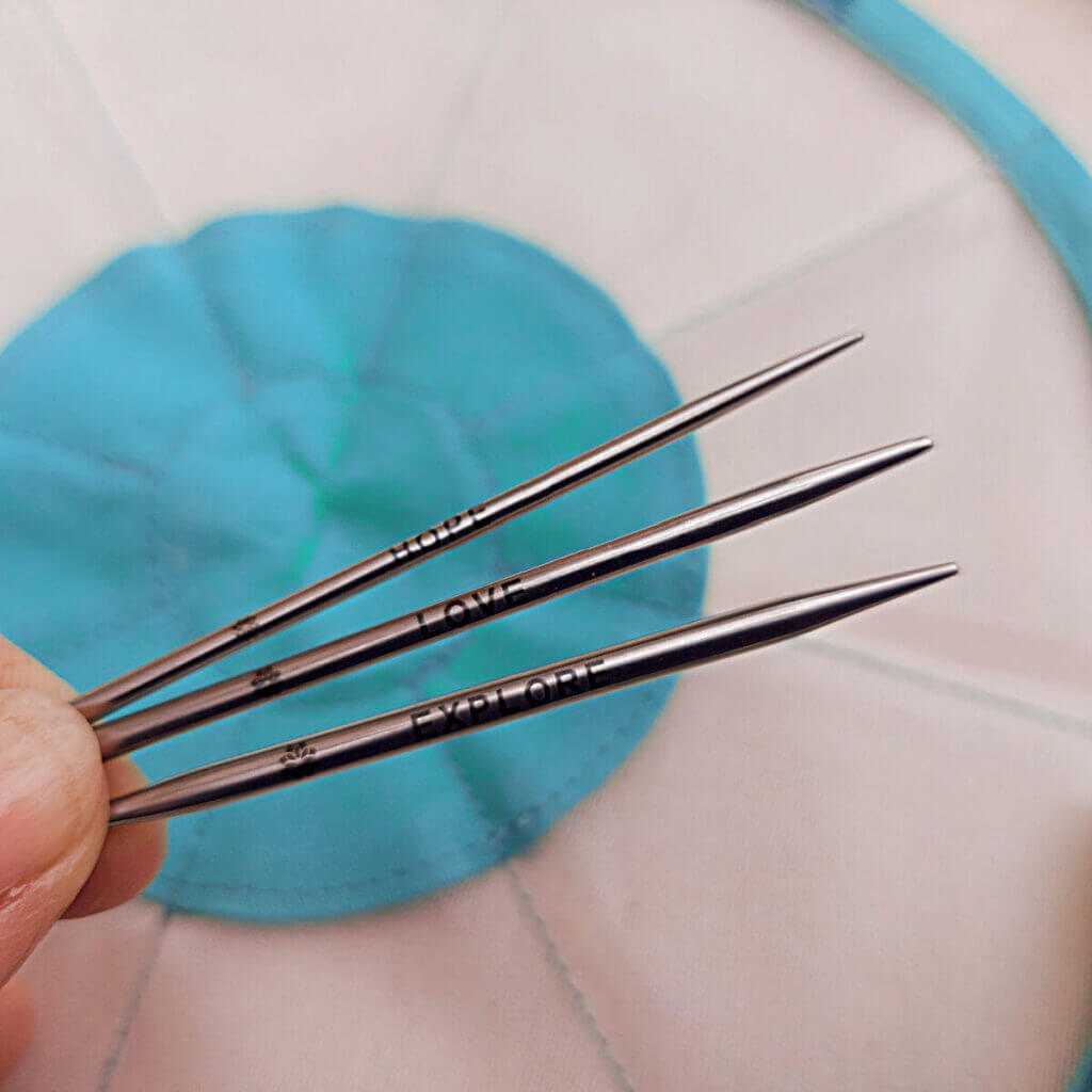 Three metal knitting needle tips, each with a word stamped on them. From the top, the words are "hope", "love", "explore"