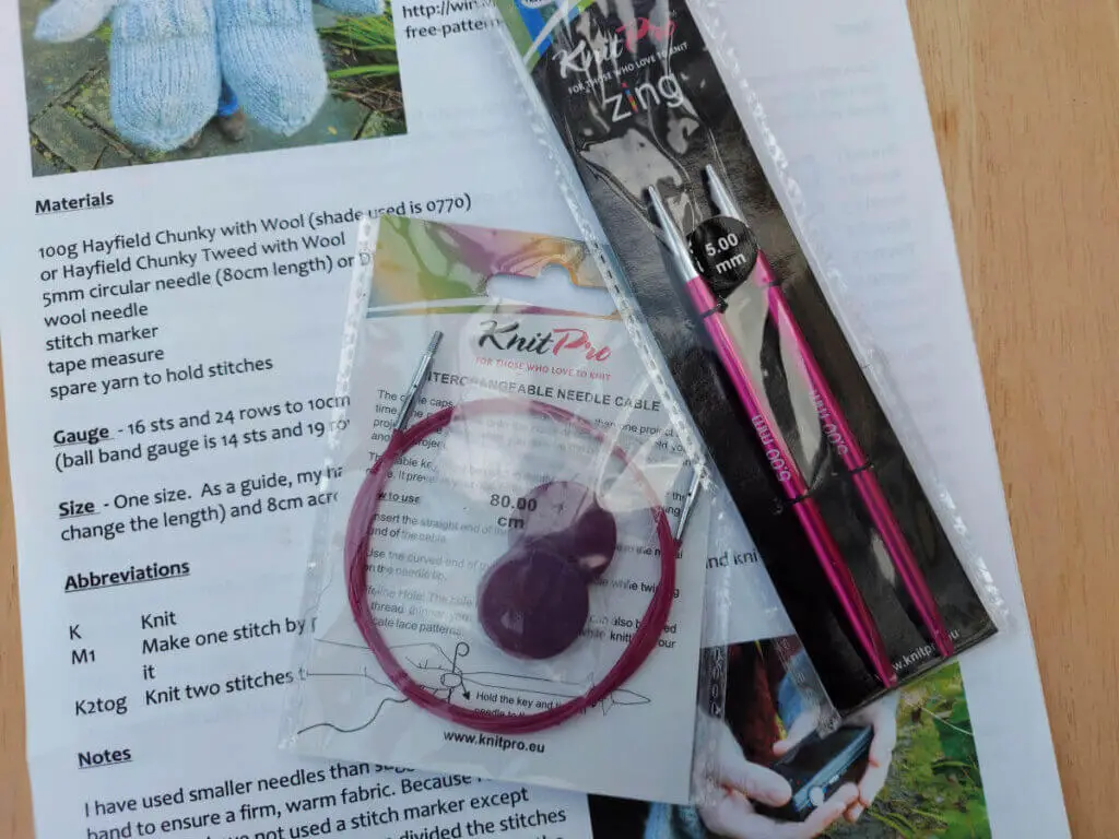 Pink needle tips in a packet next to a pink circular needle cable in another packet. They are both lying on a knitting pattern