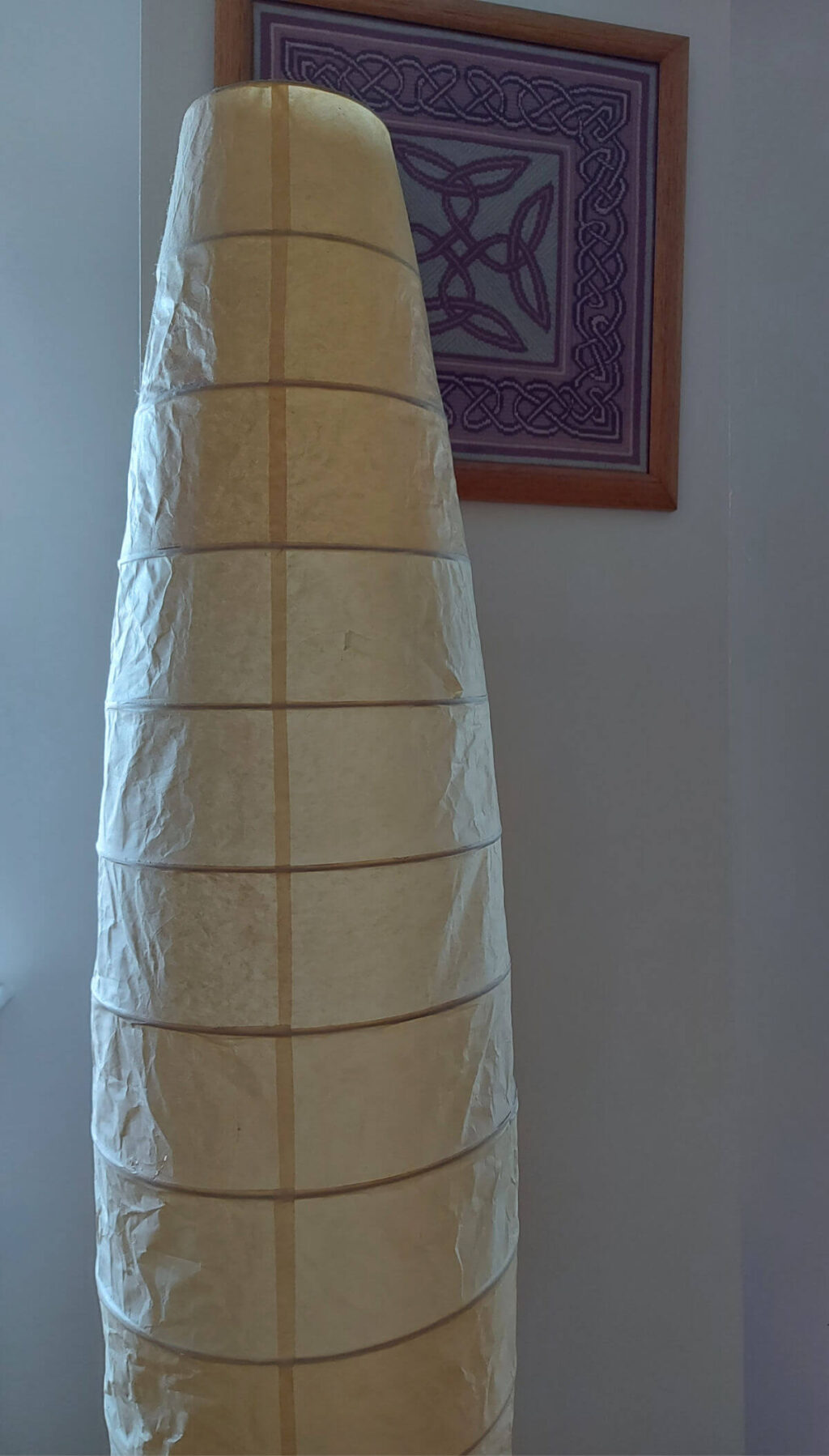 A tall lampshade with a yellowed paper shade stands against a cream wall with a pink Celtic knot tapestry on it
