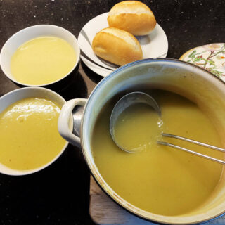 Yellowy-green leek and potato soup in a pan with a metal soup ladle resting against the side. To one side of the pan are two bowls of the soup and a small plate with two crusty bread rolls on it.
