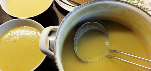 Yellowy-green leek and potato soup in a pan with a metal soup ladle resting against the side. To one side of the pan are two bowls of the soup and a small plate with two crusty bread rolls on it.