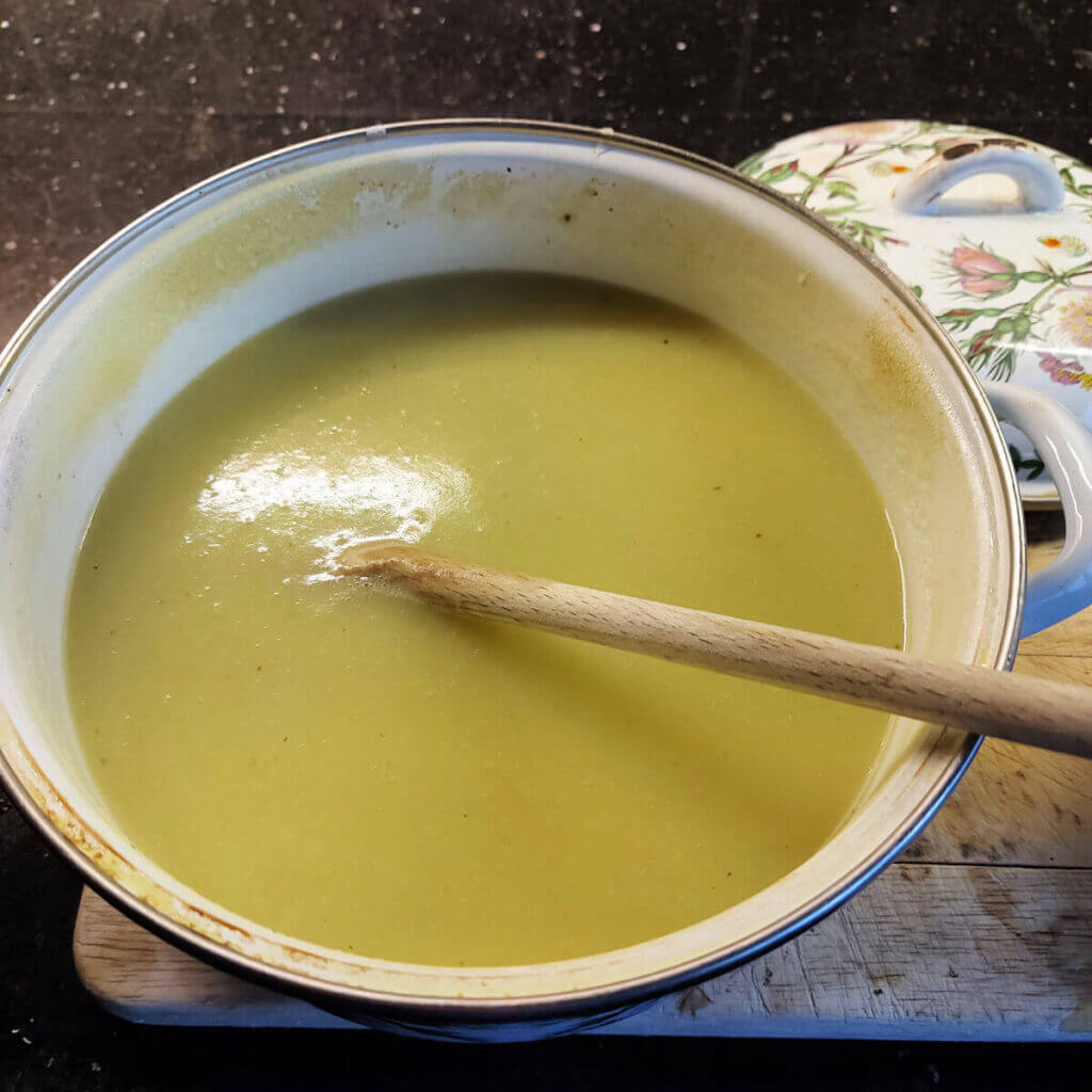 Leek and potato soup in a pan with a wooden spoon resting against the side of the pan. The soup is a yellowy-green colour.