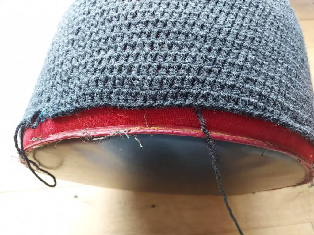 A view of the side of a red pouffe showing how much more of a grey crocheted cover needs to be worked.