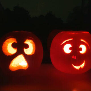 Two pumpkins with faces carved into them and lit up with candles. They are sitting on a windowsill and there are silhouettes of trees in the darkness behind them.