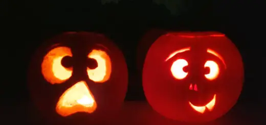 Two pumpkins with faces carved into them and lit up with candles. They are sitting on a windowsill and there are silhouettes of trees in the darkness behind them.
