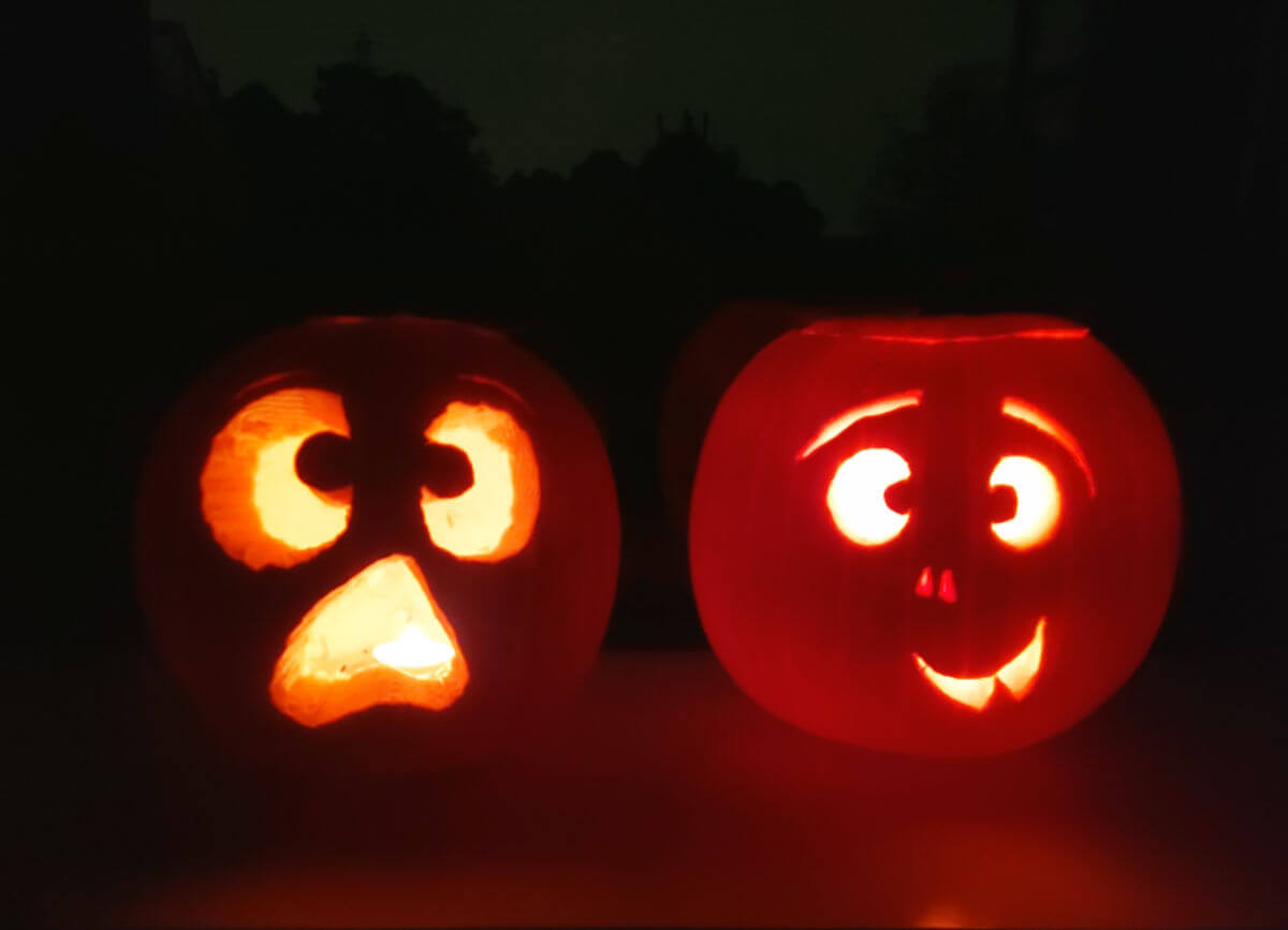 Two pumpkins with faces carved into them and lit up with candles. They are sitting on a windowsill and there are silhouettes of trees in the darkness behind them.
