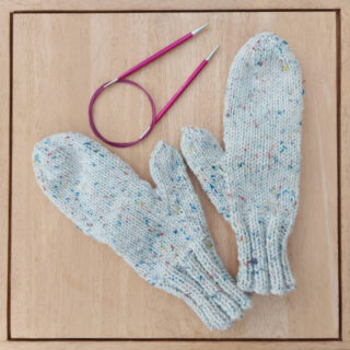 A pair of chunky mittens on a wooden table next to a set of pink circular needles