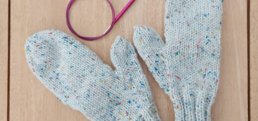 A pair of chunky mittens on a wooden table next to a set of pink circular needles