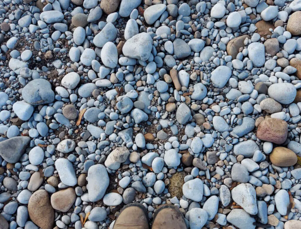 Grey and brown rounded pebbles on a river beach, Christine's boots are in the photo for context
