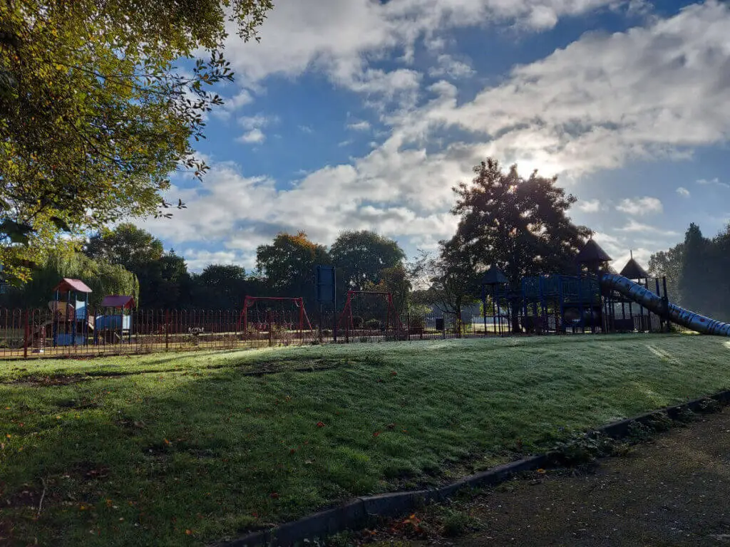 An empty playpark in the early morning sun. The sky is blue and cloudy and the sun is shining through the trees.