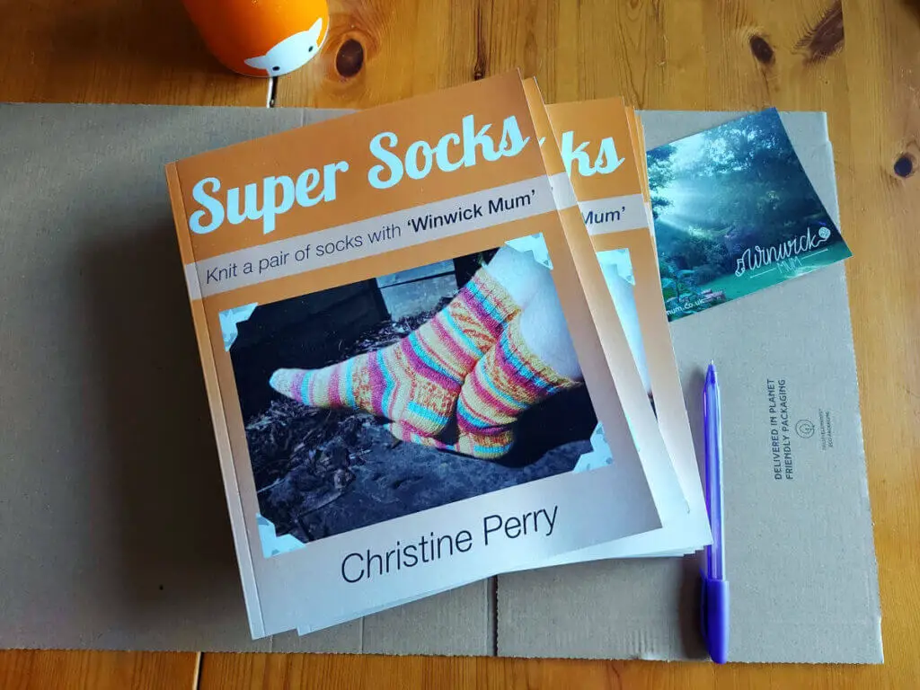 A small pile of orange Super Socks books on cardboard packaging next to a Winwick Mum postcard, a purple pen and an orange mug of tea