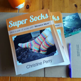 A small pile of orange Super Socks books on cardboard packaging next to a Winwick Mum postcard, a purple pen and an orange mug of tea