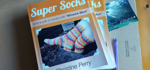 A small pile of orange Super Socks books on cardboard packaging next to a Winwick Mum postcard, a purple pen and an orange mug of tea