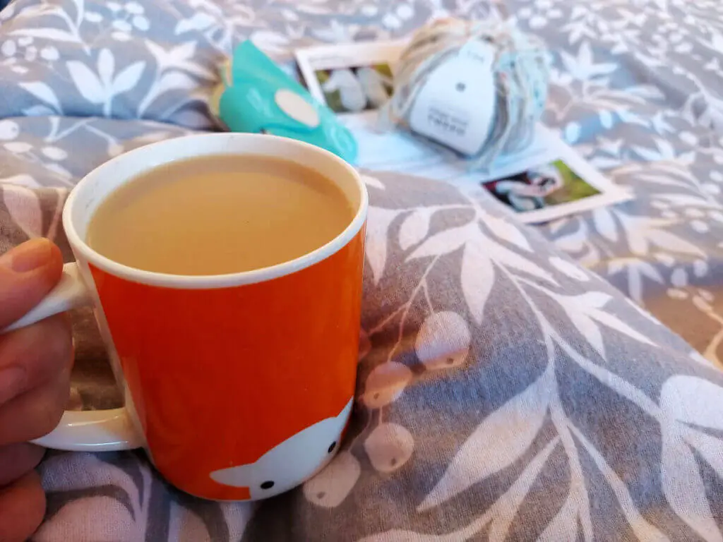 An orange mug of tea balanced on a grey and white patterned duvet cover. In the background are a knitting pattern, a ball of yarn and a blue packet of needles