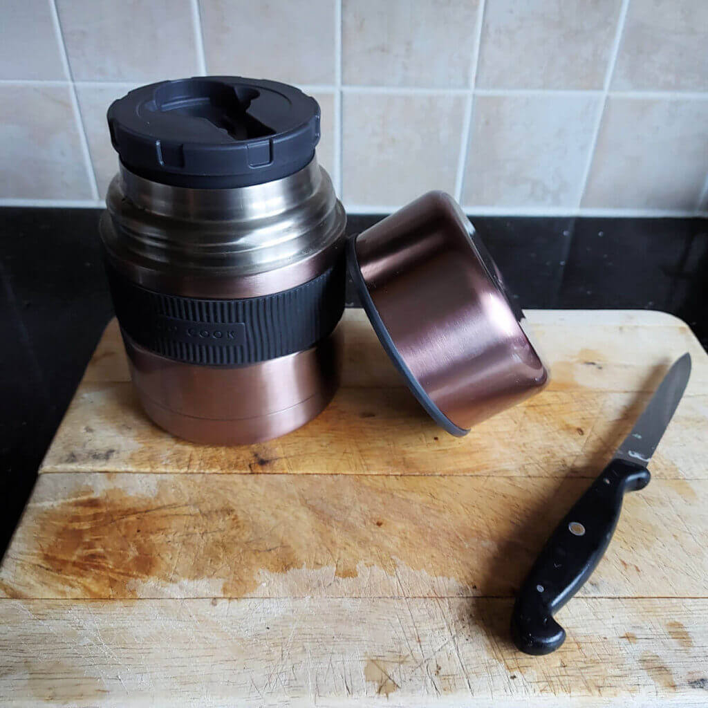 A wide-necked rose gold thermal food flask stands on a wooden chopping board next to a knife with a black handle. The worktop behind is black and the tiles on the wall are beige squares
