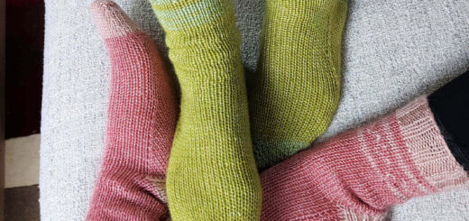 Two pairs of feet intertwined on a grey sofa. One pair of feet is wearing green socks and the other is wearing pink socks