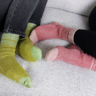 Two pairs of feet intertwined on a grey sofa. One pair of feet is wearing green socks and the other is wearing pink socks