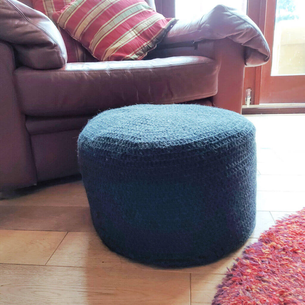 A round pouffe with a grey crocheted cover sits on a wooden floor next to a red rug and a brown leather sofa with red and gold striped cushions