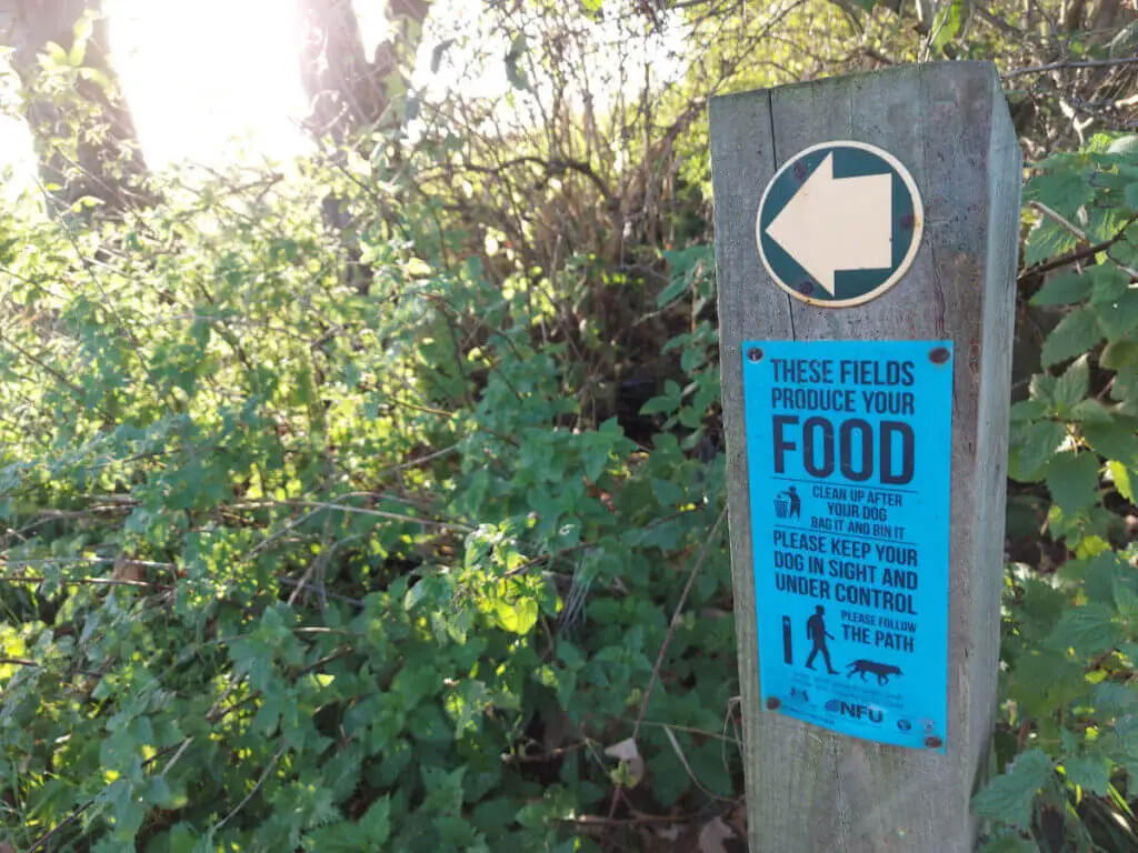 A blue sign on a wooden stake surrounded by bushes. The sign reminds footpath users that crops are grown in the fields