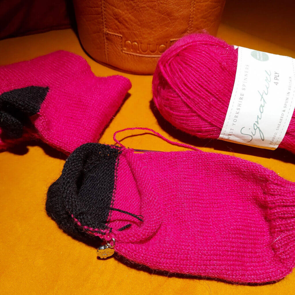 A partly-knitted pink sock with a black heel on a mustard-coloured sofa. Next to it is a completed sock, a ball of pink yarn and a brown leather project bag