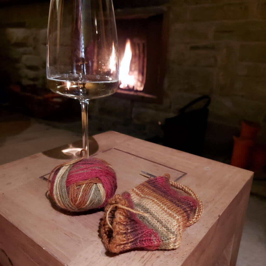 A glass of wine and partly-knitted sock in shades of red, green and brown sit on a wooden coffee table. In the background is an open fire.